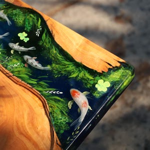 May include: Wooden table corner with a resin inlay depicting a koi pond. The design features koi carp, aquatic plants, and a dark blue water effect. The wood has a natural grain and a warm, brown tone.