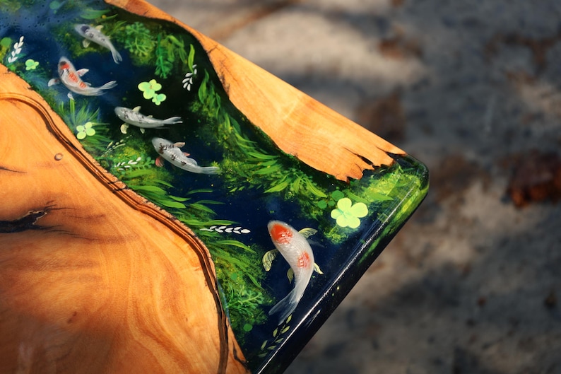 May include: Wooden table corner with a resin inlay depicting a koi pond. The design features koi carp, aquatic plants, and a dark blue water effect. The wood has a natural grain and a warm, brown tone.