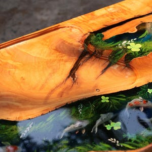 May include: A handcrafted wooden table with a unique design. The table features a live edge, showcasing the natural wood grain. The centre is filled with a clear resin, depicting a pond scene with koi fish and aquatic plants. The wood is a warm, golden brown.