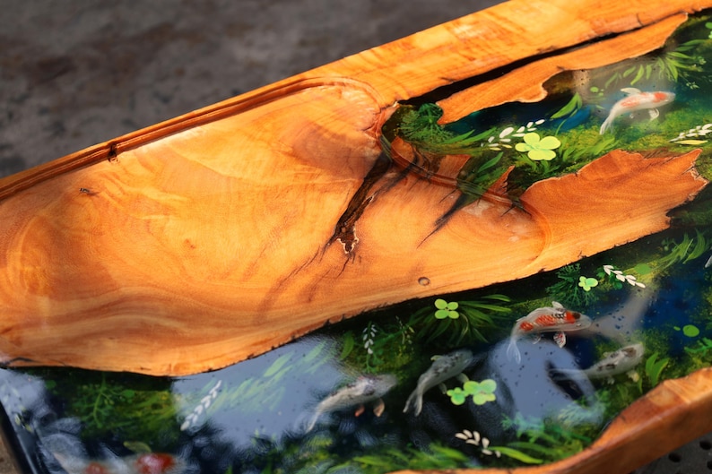 May include: A handcrafted wooden table with a unique design. The table features a live edge, showcasing the natural wood grain. The centre is filled with a clear resin, depicting a pond scene with koi fish and aquatic plants. The wood is a warm, golden brown.