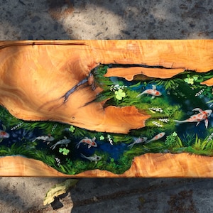 May include: A rectangular wooden serving board with a river design featuring koi carp. The wood has a natural grain with a light brown colour. The river is filled with green plants and blue water, with several koi carp swimming.