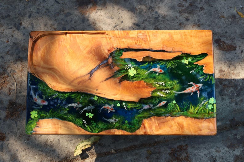 May include: A rectangular wooden serving board with a river design featuring koi carp. The wood has a natural grain with a light brown colour. The river is filled with green plants and blue water, with several koi carp swimming.