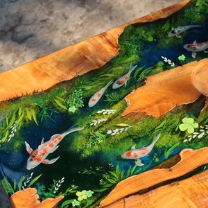 May include: A wooden table with a river scene. The table features a clear resin river with koi carp swimming among green plants. The wood has a natural, light brown colour, and the resin river is a deep blue.