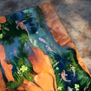 May include: A wooden table featuring a river scene with koi carp. The river is a deep blue, with the fish swimming amongst green plants. The table's edges are natural wood, creating a striking contrast.