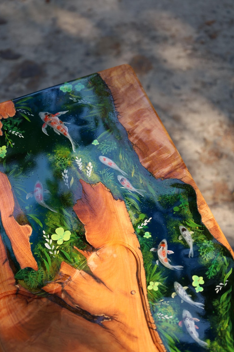 May include: A wooden table featuring a river scene with koi carp. The river is a deep blue, with the fish swimming amongst green plants. The table's edges are natural wood, creating a striking contrast.