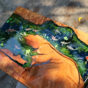 May include: A rectangular wooden serving board featuring a river scene. The river is crafted from blue resin, populated with koi carp. Green plants and yellow flowers border the river. The wood displays a natural grain pattern.