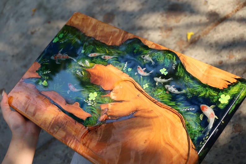 May include: A rectangular wooden serving board featuring a river scene. The river is crafted from blue resin, populated with koi carp. Green plants and yellow flowers border the river. The wood displays a natural grain pattern.