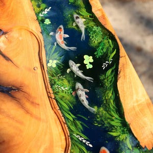 May include: A wooden table with a river of blue resin running through it. The resin river features painted koi carp and green aquatic plants. The table's wooden surface has a natural wood grain pattern.