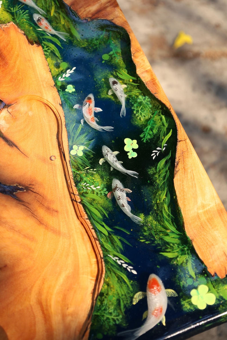 May include: A wooden table with a river of blue resin running through it. The resin river features painted koi carp and green aquatic plants. The table's wooden surface has a natural wood grain pattern.