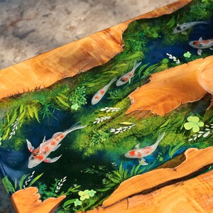 May include: A handcrafted wooden table with a river design. The river features a blue epoxy resin with koi carp and green plants. The table's edges are natural wood, showcasing the grain and texture. A unique piece for home decor.