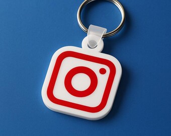 May include: White and red Instagram logo keychain with a silver keyring. The keychain features the Instagram logo in red on a white square background. The background is a solid blue.