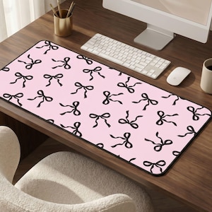 May include: A rectangular pink desk mat with a black border and a repeating pattern of black bows. A computer keyboard, mouse, and monitor are on the desk. A gold pencil holder and a mug are also visible.