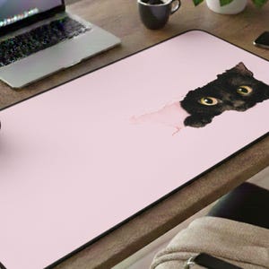 May include: A rectangular pink mouse pad with a black border. The pad features a black cat peeking through a torn section, with yellow eyes. The design is a whimsical addition to any desk setup.