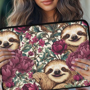 May include: A laptop sleeve featuring a whimsical design with smiling sloths and vibrant floral elements. The background is a cream color with burgundy peonies, pink flowers, and green leaves. The sloths have brown fur and are smiling.