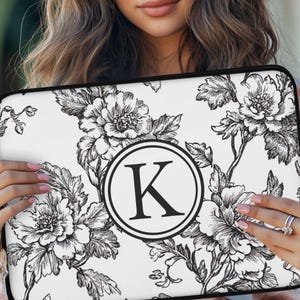 May include: A rectangular laptop sleeve featuring a white background with a black ink-style floral pattern. Centered on the sleeve is a black circular emblem containing the capital letter K in a serif font, all enclosed by a thin black border.
