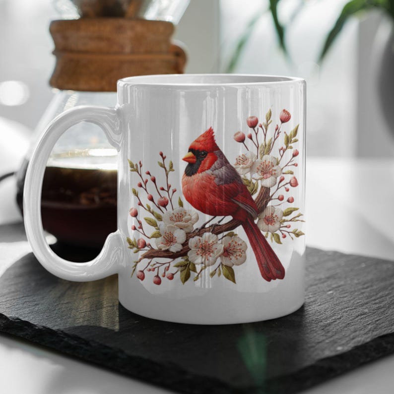 May include: White ceramic mug featuring a red cardinal perched on a branch of white blossoms. The cardinal has a black eye and a long, red tail. The flowers are surrounded by green leaves.