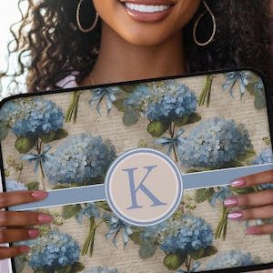 May include: Laptop sleeve featuring a floral pattern of blue hydrangeas and ribbons on a beige background with script. A light blue band and a circular emblem with the letter "K" are centered on the sleeve.