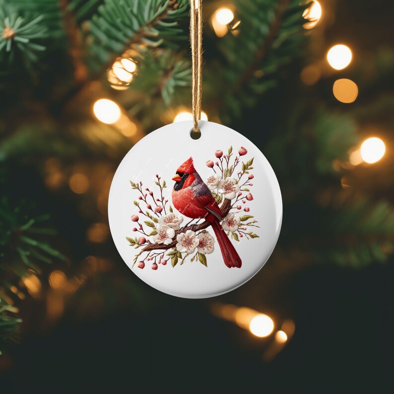 May include: A white ceramic bauble featuring a red cardinal perched on a branch with white blossoms. The cardinal is surrounded by green leaves and pink buds.