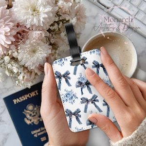 May include: White luggage tag with a blue bow and floral pattern. The tag has a black leather strap and is held in a person's hand. The tag is decorated with blue bows and floral bouquets. The text "Monarch Artistry & Design" is visible.