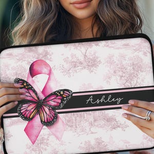 May include: A pink and black laptop sleeve featuring a pink ribbon and a butterfly design. The sleeve has a black stripe with the name "Ashley" in white script. The background has a pink and white toile pattern.