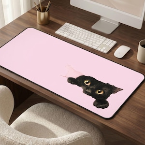 May include: A pink rectangular desk mat with a black border. A black cat with yellow eyes is peeking through a torn section of the mat. A computer keyboard, mouse, and monitor are visible.