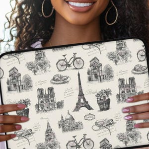 May include: A cream-colored laptop sleeve featuring a black and white Parisian theme. The design includes illustrations of the Eiffel Tower, bicycles, croissants, and other Parisian landmarks. The sleeve is held by a person.