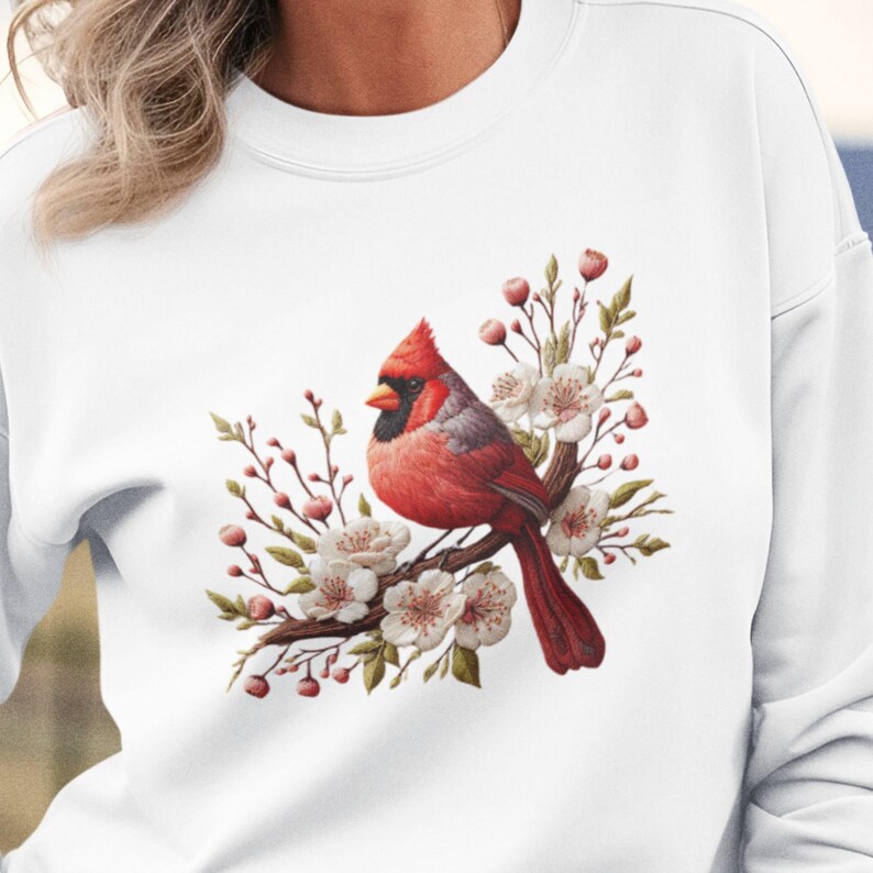 May include: A white sweatshirt featuring a red cardinal bird perched on a branch with white blossoms. The bird is surrounded by green leaves and pink buds.