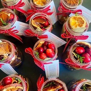 May include: Several small glass jars filled with dried orange slices, cranberries, cinnamon sticks, and bay leaves. Each jar is tied with a red and white polka dot ribbon. Small white cards with text are attached to the jars. The jars are arranged on a dark surface.