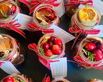 May include: Multiple glass jars containing dried orange slices, cranberries, cinnamon sticks, and bay leaves. Each jar is adorned with a red and white polka dot ribbon. Small white cards with text are attached. The jars are displayed on a dark surface.