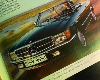 May include: A black Mercedes-Benz SL-Class convertible with a silver license plate that reads "SHV 9530". The car is driving on a road with green grass and trees in the background.