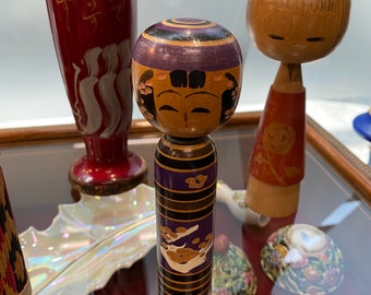 May include: A wooden Japanese doll with a purple and black painted design. The doll has a painted face with black hair and a purple headband. The doll is standing on a table with other decorative items.