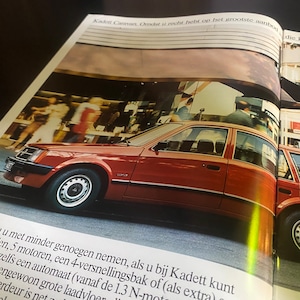 Op de afbeelding: Een rode Opel Kadett staat geparkeerd op een straat in de stad. De auto heeft een zwarte bumper en zwarte bekleding rond de ramen. De auto is scherp, terwijl de achtergrond wazig is.