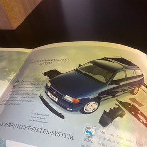 May include: A blue Opel Astra station wagon with the text "DAS ASTRA-RECYCLING-SYSTEM" and "DAS ASTRA-REINLUFT-FILTER-SYSTEM" on the page. The car is surrounded by various car parts.