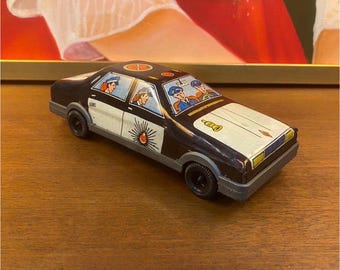 Könnte beinhalten: Ein altes Polizeiauto-Spielzeug aus Blech in Schwarz und Weiß mit grauer Stoßstange. Das Auto zeigt Illustrationen von Beamten in den Fenstern, eine Sirenen-Grafik an der Seitentür und ein Hupen-Symbol auf der weißen Motorhaube.