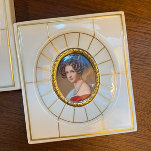Op de afbeelding: Een wit porseleinen fotolijst met een gouden rand en een portret van een vrouw in een rode jurk. De lijst heeft een decoratief ontwerp met gouden lijnen.