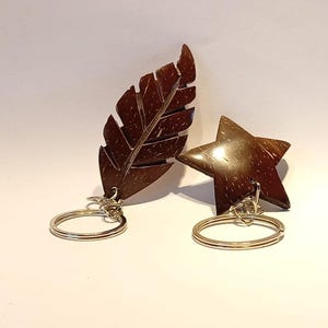 Pode incluir: Dois porta-chaves feitos de material de casca de coco polido castanho escuro. Um porta-chaves tem a forma de uma folha detalhada com bordas serrilhadas e o outro tem a forma de uma estrela de cinco pontas. Ambos possuem argolas de metal prateado.