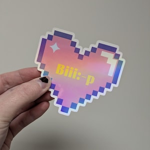 May include: A pixelated heart-shaped sticker with a holographic finish. The sticker features a gradient of pink, yellow, and blue hues. The text "Biii:-p" is printed in yellow across the center of the heart. The sticker has a white border and a pixelated design.