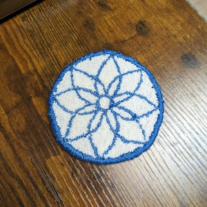 May include: A blue and white embroidered coaster with a floral design. The coaster is round and has a blue border.