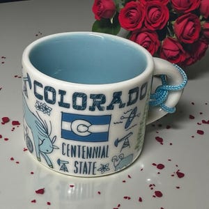 May include: A white ceramic mug with a light blue interior and a light blue cord tied to the handle. The exterior features blue illustrations including the text Colorado Centennial State, the Colorado state flag, a bighorn sheep, and oil derricks.