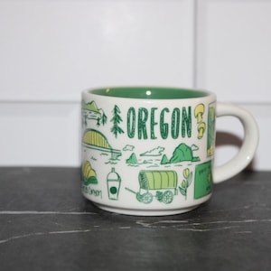 May include: White ceramic mug with a green interior, featuring green and yellow illustrations of Oregon landmarks and the word "OREGON". The mug has a handle and is set against a white tile background.