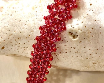 May include: A red beaded bracelet with a bronze clasp. The bracelet is made of small, round beads that are strung together in a pattern. The clasp is a simple, round ring.