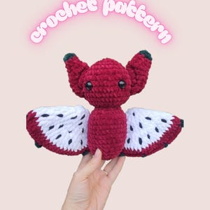 May include: A handmade crochet bat with a red body and wings designed to resemble dragon fruit. The wings are white with black speckles and red trim. The text "crochet pattern" is above the bat.