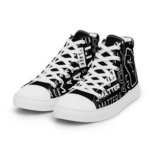 May include: Black and white high-top sneakers with white laces. The sneakers feature the words "Black Lives Matter" in a repeating pattern, along with a raised fist graphic. The soles and toe caps are white.