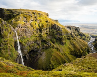 May include: A cascading waterfall plunges down a steep, moss-covered cliff face in a rugged, green landscape. A winding river flows through the valley below, leading to a distant, flat plain.