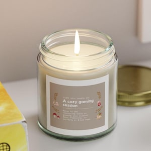 May include: A glass candle jar with a beige label that reads "Take this candle for A cozy gaming session". The candle is lit and the jar is sitting on a white shelf with a book and a potted succulent plant.
