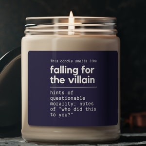 May include: A lit candle in a clear glass jar with a black lid. The candle has a white wax and a dark blue label with the text "falling for the villain". The text also includes "hints of questionable morality; notes of 'who did this to you?'".