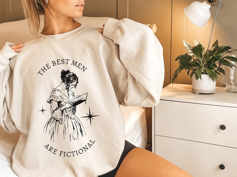 May include: A light beige sweatshirt with a black graphic print of a woman reading a book. The text on the sweatshirt reads "The Best Men Are Fictional."