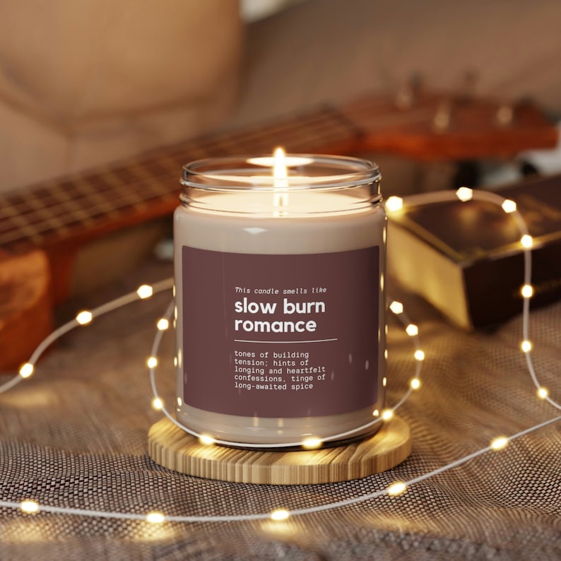 Puede incluir: Un tarro de vidrio con una vela y una etiqueta marr&oacute;n que dice "This candle smells like slow burn romance" y una descripci&oacute;n del aroma: "tones of building tension; hints of longing and heartfelt confessions, tinge of long-awaited spice".