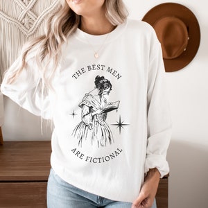 May include: White long-sleeve sweatshirt with a black and white illustration of a woman reading a book. The text "THE BEST MEN ARE FICTIONAL" is printed on the shirt.
