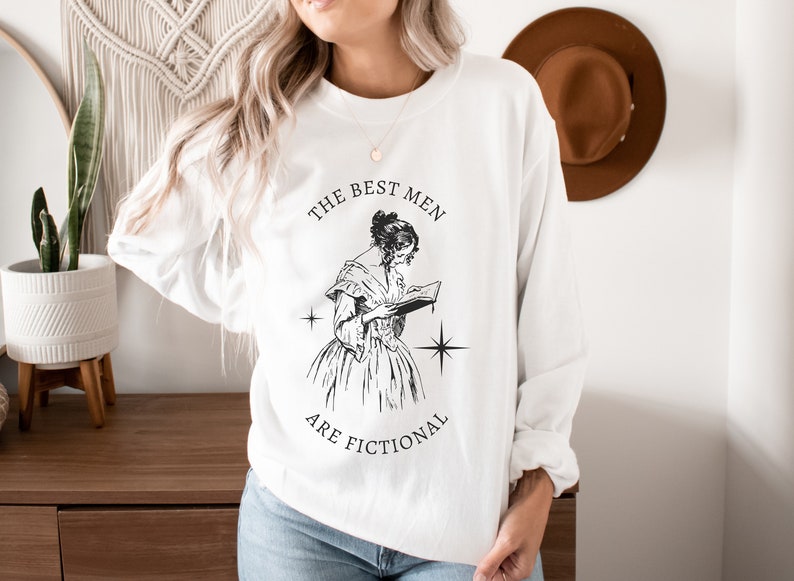 May include: White long-sleeve sweatshirt with a black and white illustration of a woman reading a book. The text "THE BEST MEN ARE FICTIONAL" is printed on the shirt.